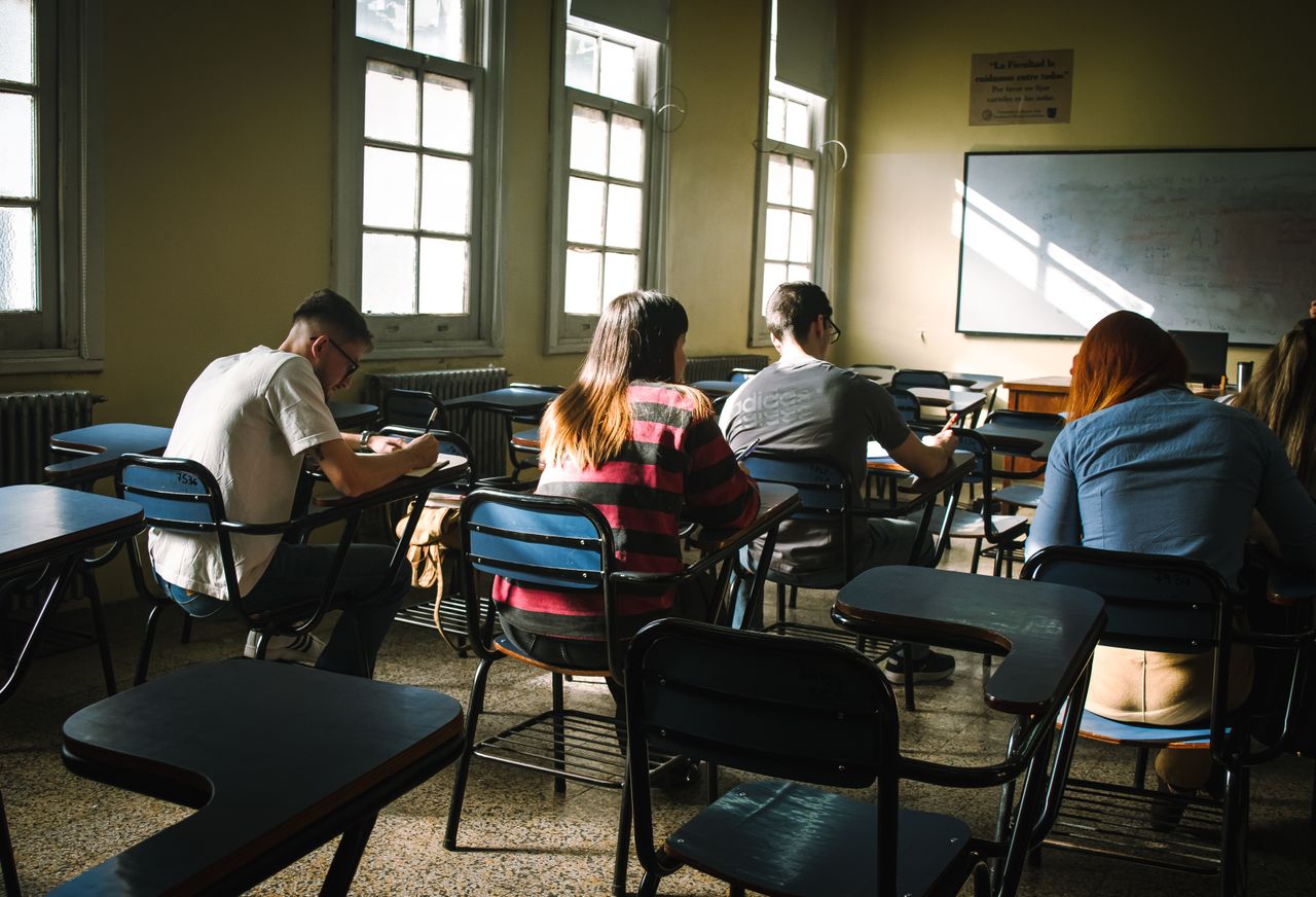 A group of students focused on writing in a sunlit classroom with desks and windows.